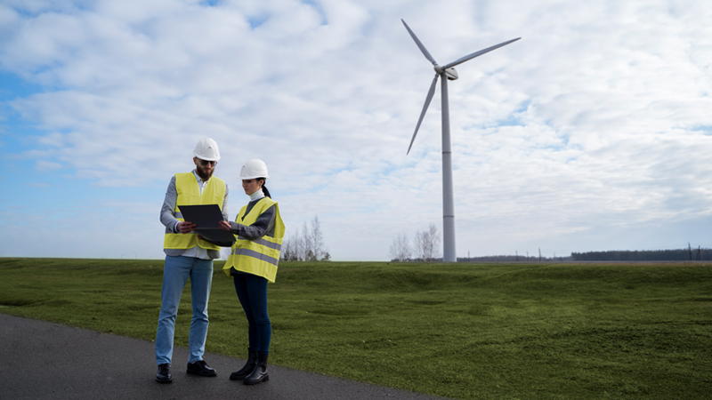 Man and woman workers with laptop, standing in front of a wind turbine.